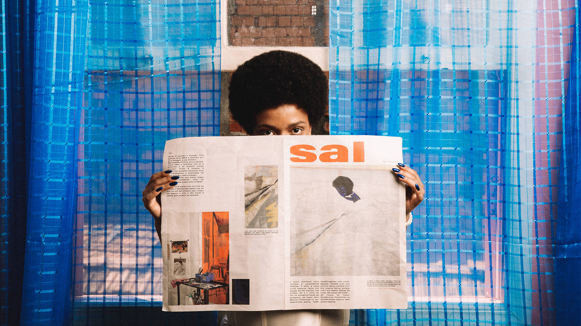 Anelis segura um jornal, em uma das p&aacute;ginas est&aacute; a capa do seu &aacute;lbum mais recente, Sal.