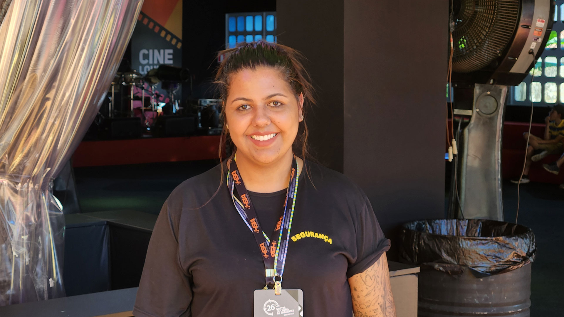 Eloa sorri na foto. Veste um crach&aacute;. Atr&aacute;s dela &eacute; poss&iacute;vel ver um espa&ccedil;o com janelas e palco.