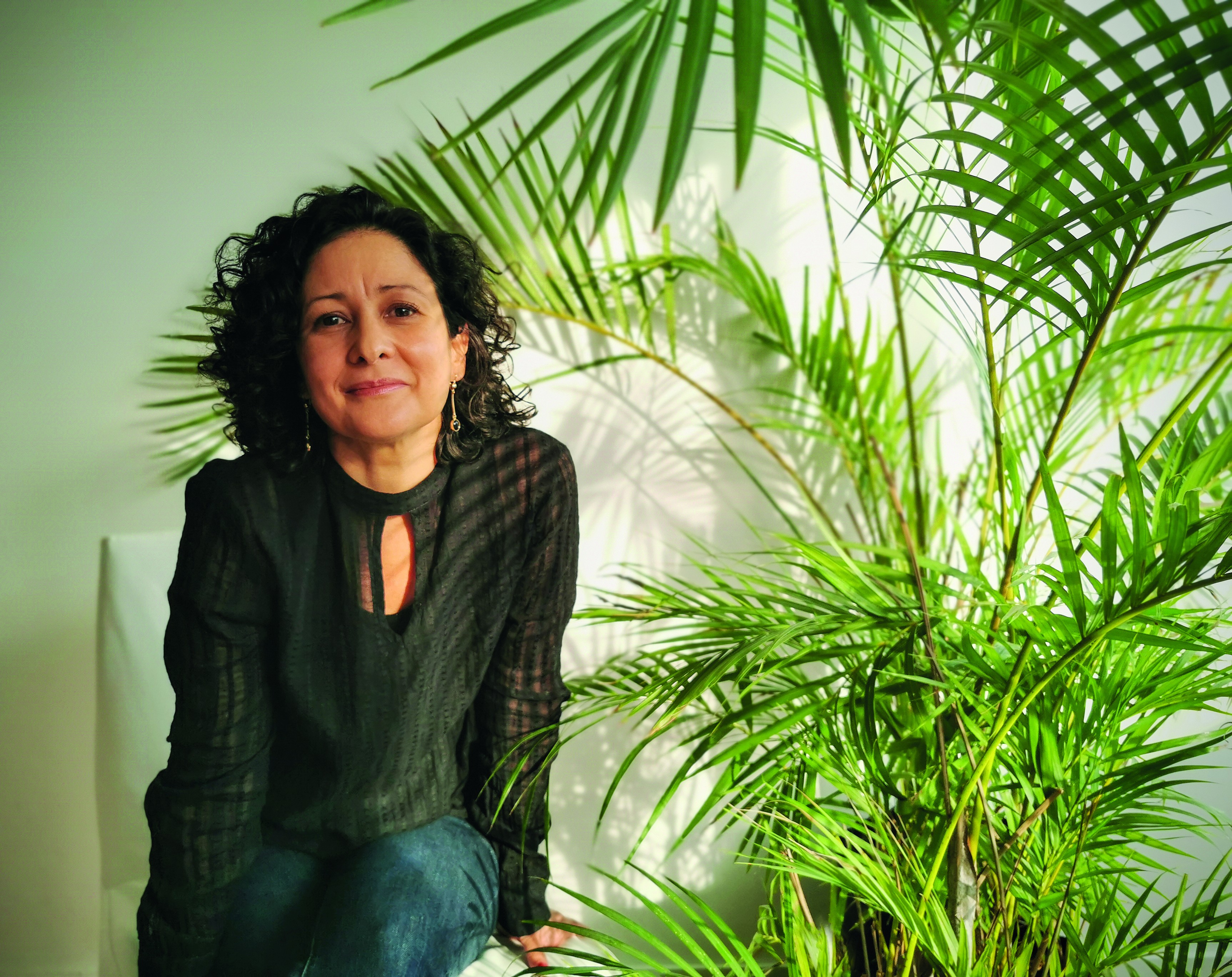 Mulher de pele morena e cabelos encaracolados escuros, sorrindo levemente, sentada em uma cadeira branca, vestindo blusa preta e cal&ccedil;a jeans, ao lado de uma planta verde exuberante com folhas longas e finas, contra uma parede clara