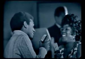 Dois cantores negros, um homem e uma mulher, sorriem enquanto seguram microfones em um palco escuro, com um homem de terno ao fundo