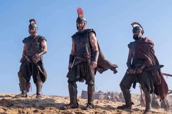 Três homens com armaduras e capacetes antigos, um com penacho vermelho, estão em uma paisagem desértica com tendas ao fundo, sob céu azul