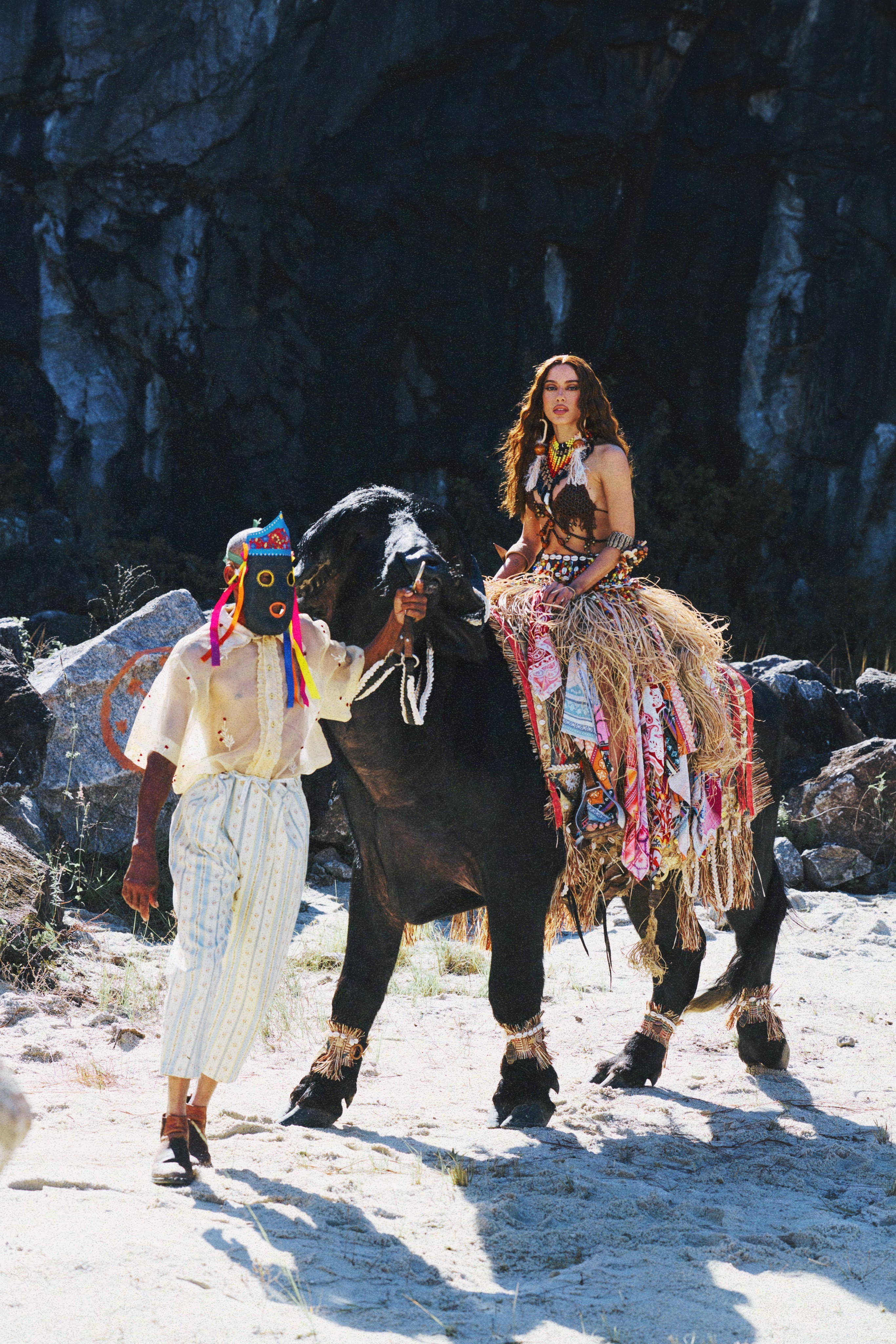 Mulher com cabelos longos e ondulados, usando top e saia de palha e mi&ccedil;angas, sentada em um b&uacute;falo escuro ricamente decorado. Um homem com m&aacute;scara colorida e roupas claras guia o animal. O cen&aacute;rio &eacute; um terreno arenoso com rochas e vegeta&ccedil;&atilde;o seca, sob luz solar intensa