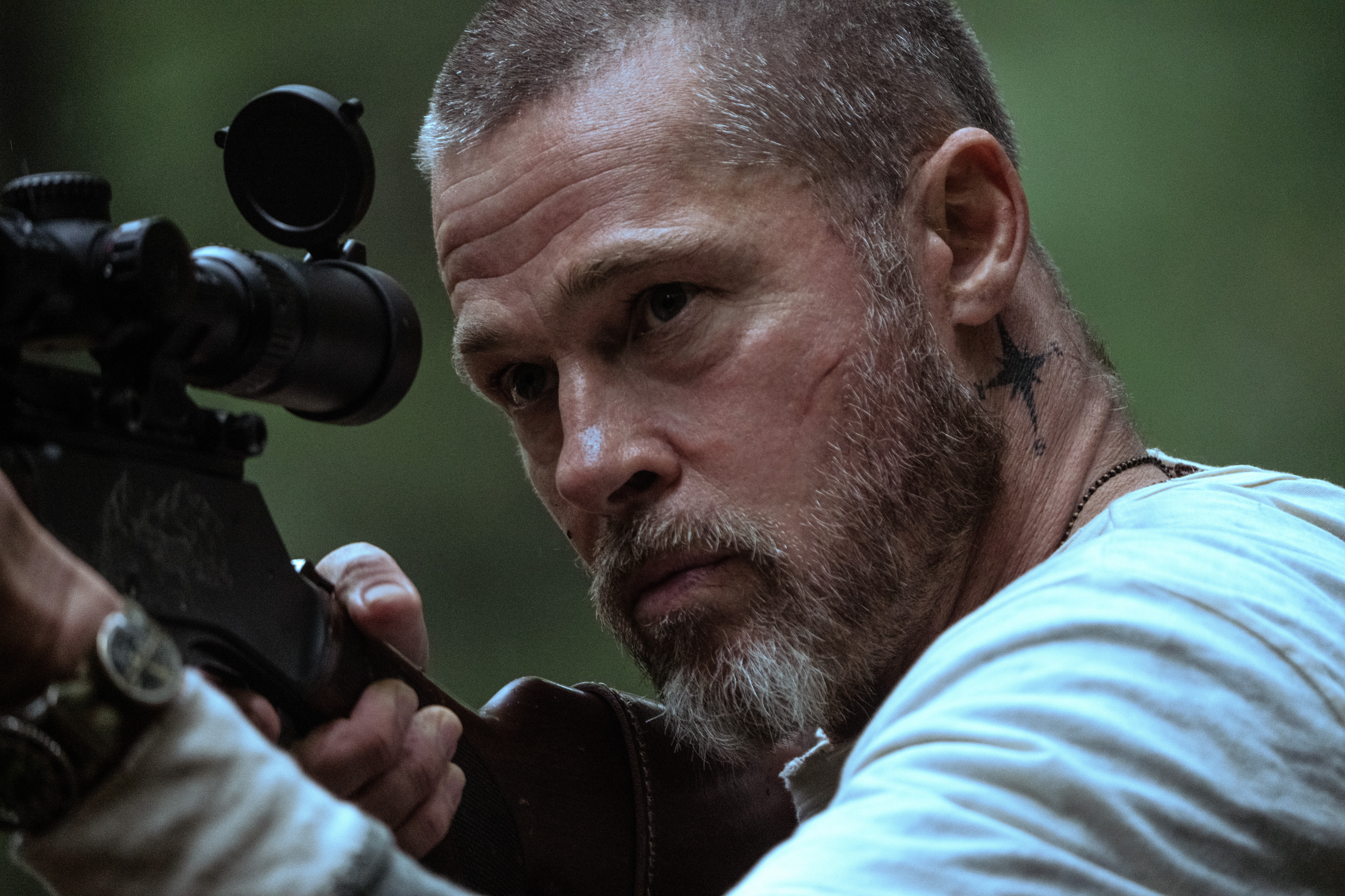 Homem de barba grisalha e cabelo curto, com cicatrizes no rosto e tatuagem de estrela no pescoço, mira com um rifle de precisão em uma floresta, usando uma camiseta branca e expressão séria