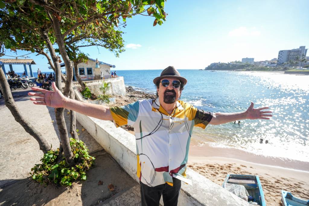 Homem de barba e chap&eacute;u, com camisa estampada, &oacute;culos escuros, bra&ccedil;os abertos, sorrindo para a c&acirc;mera em uma praia ensolarada com mar azul e barcos na areia