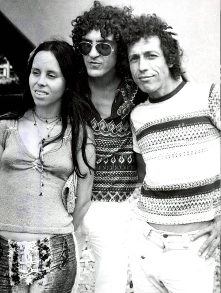 Tr&ecirc;s pessoas, uma mulher e dois homens, posam em preto e branco. A mulher &agrave; esquerda usa blusa clara, cal&ccedil;a jeans e colares. O homem do meio, de &oacute;culos escuros e cabelo cacheado, veste camisa estampada. O homem &agrave; direita, tamb&eacute;m de cabelo cacheado, usa camiseta listrada e cal&ccedil;a clara.